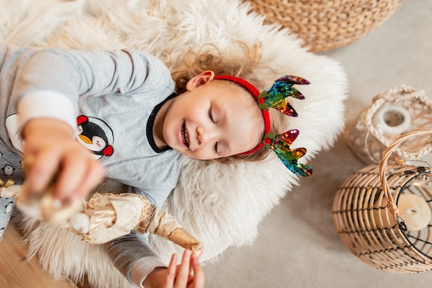 Niña feliz con asta de ciervo de Navidad en pijama de moda se encuentra en la cama y juega con un juguete en vacaciones de invierno, vistas superiores