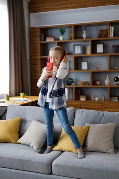 Niña feliz con auriculares rojos bailando y cantando usando un teléfono inteligente como micrófono Niño lindo divirtiéndose saltando en el autocar Ocio en casa niño feliz Trastorno por déficit de atención con hiperactividad