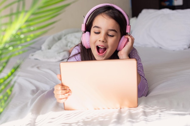 Niña feliz con auriculares rosas acostada en una cama blanca sosteniendo una tableta digital rosa