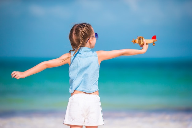 Niña feliz con avión de juguete en manos en la playa de arena blanca.