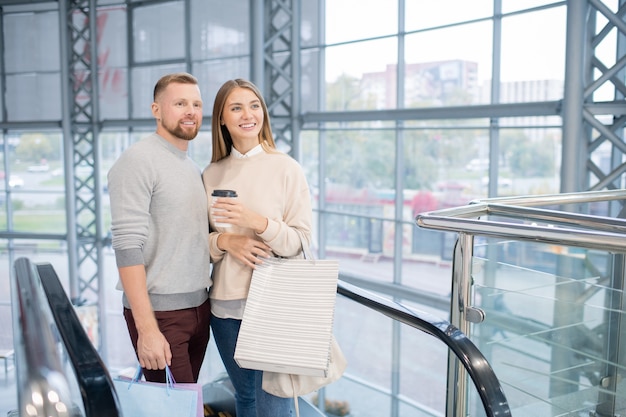 Niña feliz con bebida y bolsas y su novio mirando uno de los departamentos en el centro comercial mientras está parado en la escalera mecánica