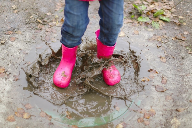 Niña feliz con botas de goma de chaqueta impermeable rosa salta alegremente a través de charcos en la calle en clima lluvioso Primavera otoño Diversión infantil al aire libre después de la lluvia Recreación al aire libre