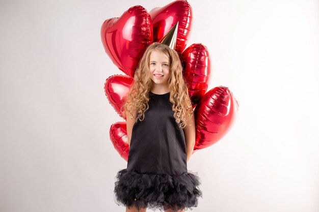 Niña feliz con brazada de bolas en forma de corazones y una gorra para su cumpleaños.