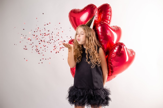 Niña feliz con brazada de bolas en forma de corazones y una gorra para su cumpleaños.