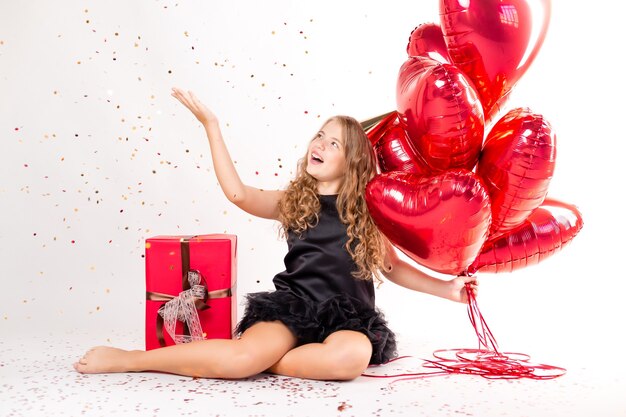 Niña feliz con brazada de bolas en forma de corazones y una gorra para su cumpleaños.