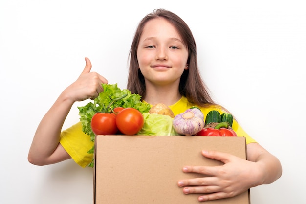 Niña feliz con una caja de verduras en las manos muestra el pulgar hacia arriba. Concepto de entrega de alimentos frescos y saludables.