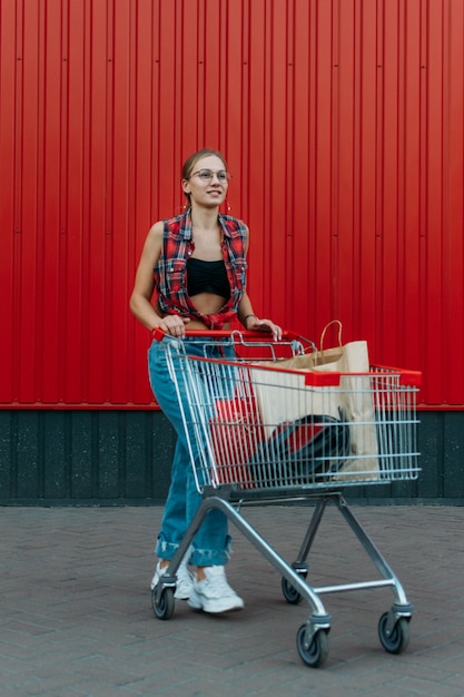 Niña feliz con carrito de compras en pared roja tienda fondo joven empujando un carrito de compras lleno