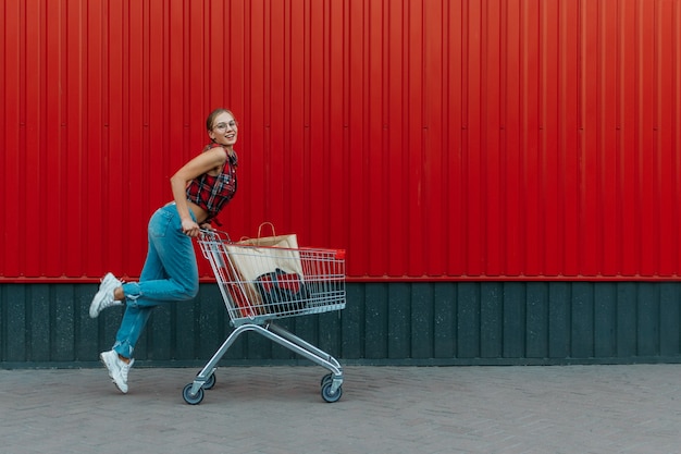 Niña feliz con carrito de compras en pared roja tienda fondo joven empujando un carrito de compras lleno