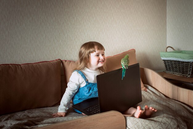 Niña feliz en casa. mascota casera. periquito. laptop y gadgets. Bebé viendo dibujos animados, juegos en línea.