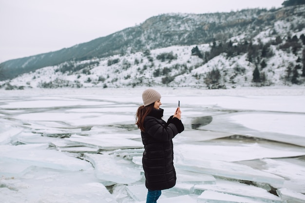 Niña feliz con una chaqueta de invierno se encuentra en el contexto de un río helado y toma la foto en un hermoso paisaje.