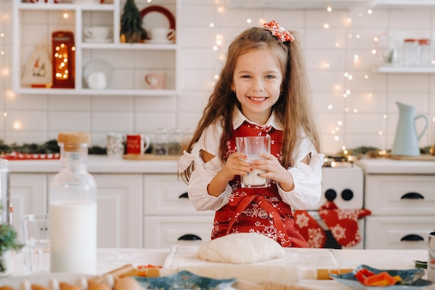 Una niña feliz en la cocina de Navidad está de pie con un vaso de leche en las manos.