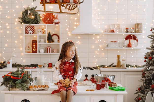 Una niña feliz en la cocina de Navidad está sentada en la mesa con galletas en sus manos.