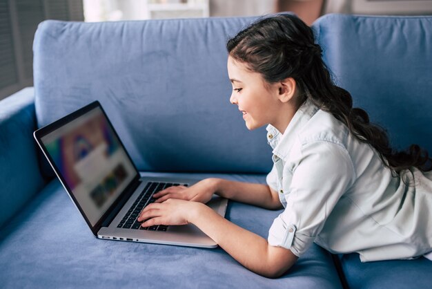 La niña feliz con una computadora portátil en el sofá