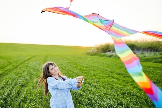 Niña feliz corriendo con kate en las manos en el campo de trigo verde Gran cometa de arco iris de colores con cola larga