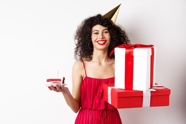 Niña feliz cumpleaños en vestido rojo, celebrando y sosteniendo regalos con pastel de cumpleaños, de pie sobre fondo blanco.