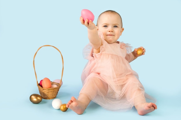 Niña feliz de diez meses con diadema de orejas de conejo sentada en un estudio azul claro y jugando con huevos de Pascua de colores Tarjeta de felicitación de Pascua para niños pequeños Conejito de Pascua para bebés