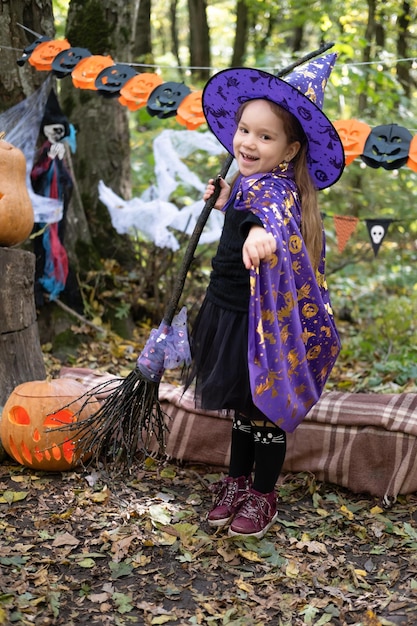 niña feliz en disfraz de halloween y sombrero de bruja con escoba en decoración de halloween al aire libre