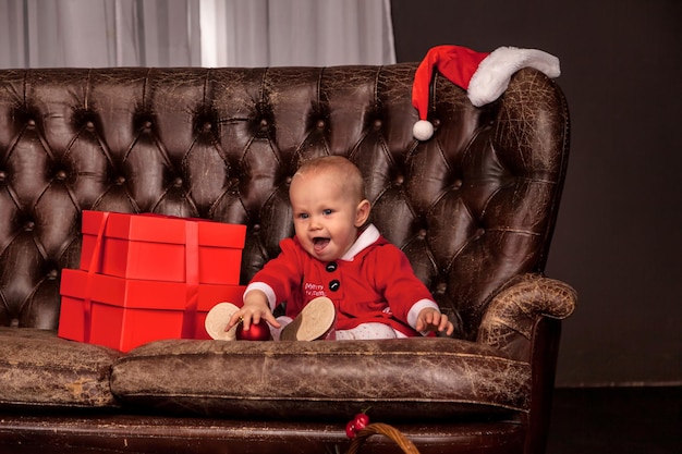 Una niña feliz disfrazada de Santa Claus sentada en un viejo sofá en el fondo decorado con un árbol de Navidad mirando a la cámara