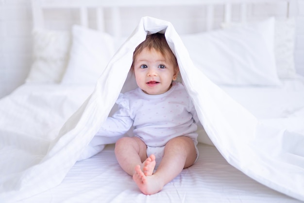 Una niña feliz y divertida mira desde debajo de la manta en la cama con ropa blanca y sonríe un niño pequeño en una cama de algodón en casa se despertó por la mañana después de dormir