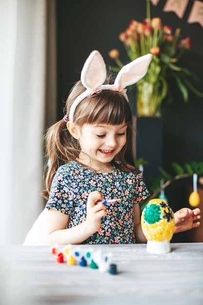 Niña feliz en edad preescolar con orejas de conejo sentada en la mesa pintando huevos de Pascua de yeso