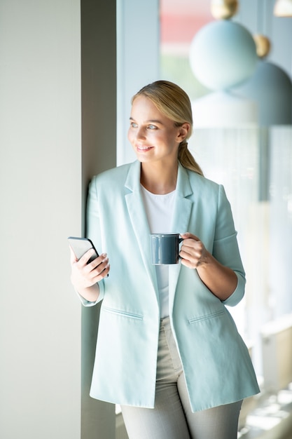 Niña feliz en elegante casual mirando en la ventana mientras se relaja en la cafetería, tomando una copa y usando el teléfono inteligente