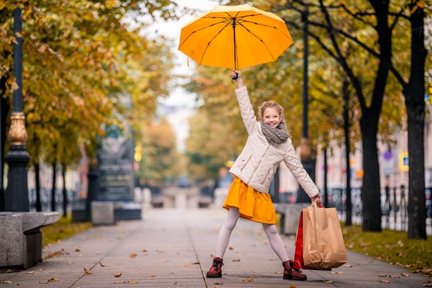 Niña feliz se encuentra en una calle de la ciudad de otoño con bolsas de la compra y un paraguas amarillo brillante.