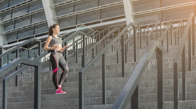 Niña feliz en el entrenamiento de cardio por la mañana. Joven mujer deportiva con smartphone eligiendo lista de reproducción para entrenamiento, sosteniendo una botella de agua