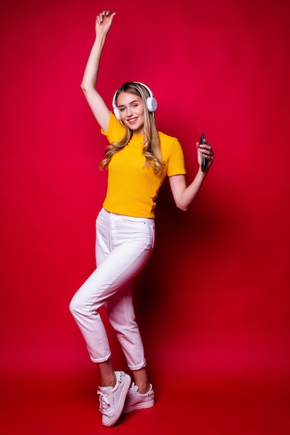 Niña feliz con, escuchando música en auriculares y bailando, sosteniendo un teléfono inteligente. En una pared roja.