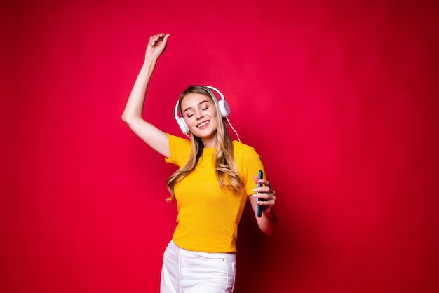 Niña feliz con, escuchando música en auriculares y bailando, sosteniendo un teléfono inteligente. En una pared roja.