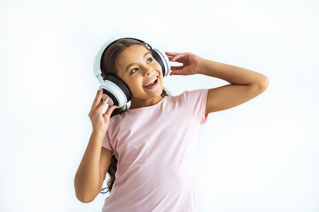 La niña feliz escuchando música en auriculares en el fondo de la pared blanca