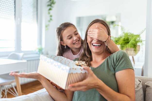 Niña feliz felicitando a la madre sonriente y dando tarjeta con corazón rojo durante la celebración navideña en casa, tapándose los ojos y sorprendiéndola