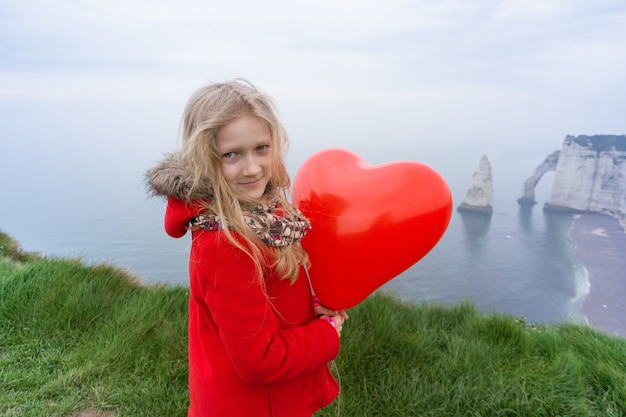 Niña feliz con un globo rojo en forma de corazón en el fondo del paisaje Etretat. Francia