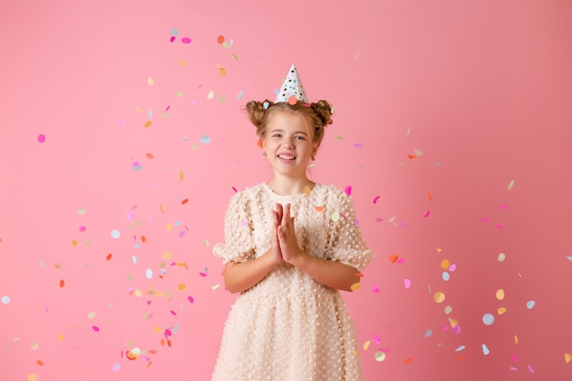 Niña feliz en una gorra de cumpleaños sopla sus palmas confeti multicolor sobre un fondo rosa en el estudio