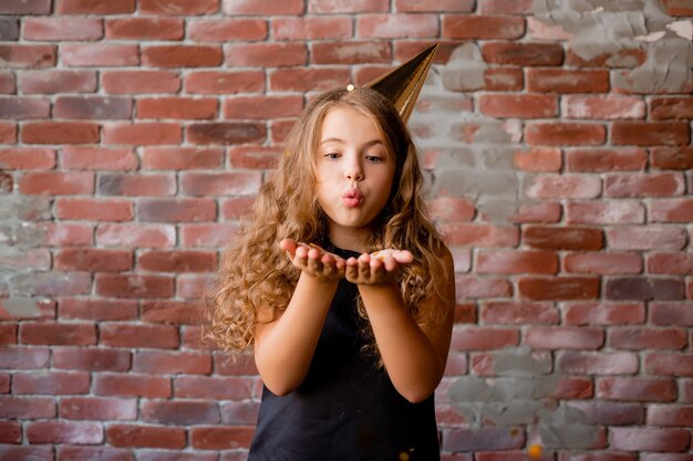 Niña feliz con una gorra para su cumpleaños sopla lejos de las palmas de confeti