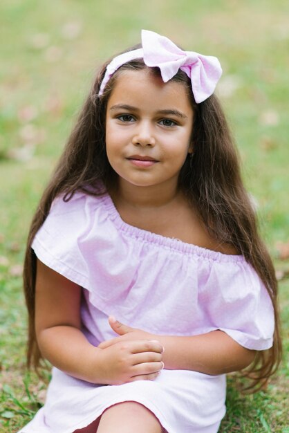 Foto niña feliz con un hermoso vestido rosa en el parque sobre el césped y mirando a la cámara