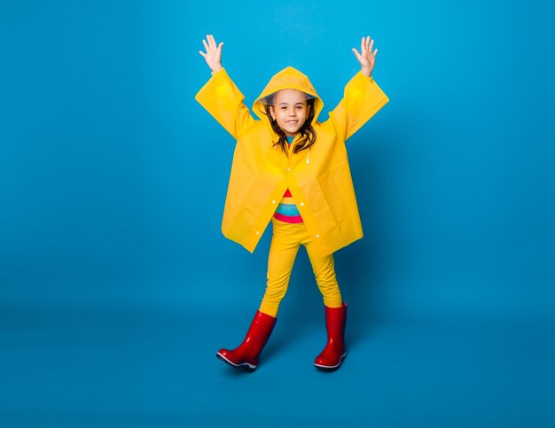 Niña feliz en un impermeable amarillo y botas rojas sobre un fondo azul.