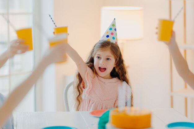 La niña feliz de jengibre usa sombreros de fiesta, tintinea tazas con bebida, se divierte con amigos, celebra cumpleaños juntos