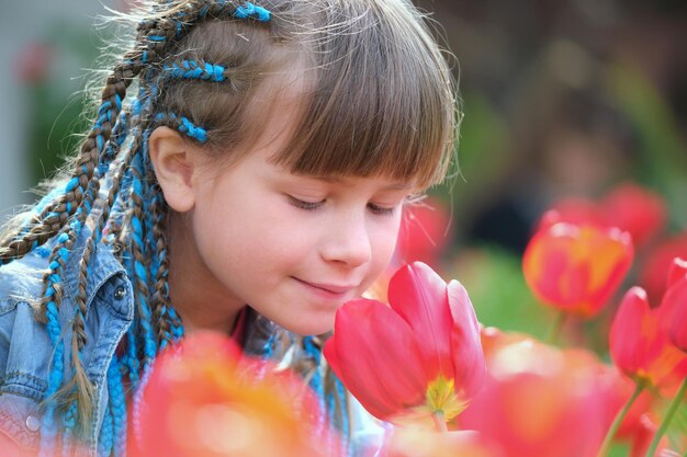 Niña feliz jugando en el jardín de verano disfrutando del dulce aroma de las flores de tulipán rojo en un día soleado