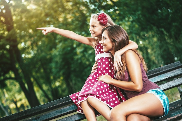 Una niña feliz y linda está jugando en el parque con su madre señalando algo. Se están abrazando y sonriendo.