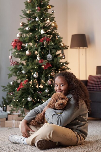 Niña feliz con linda mascota sentada en el suelo contra el árbol de Navidad