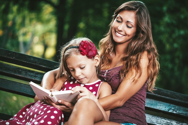 Una niña feliz y linda y su madre están leyendo un libro en un banco en el parque.