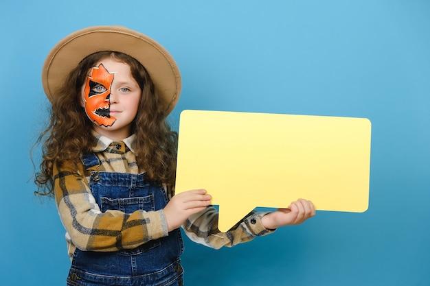 Niña feliz con máscara de maquillaje de Halloween con bocadillo amarillo, viste sombrero, positivo mirando a cámara, posando aislado sobre fondo azul de estudio con espacio de copia. Concepto de vacaciones