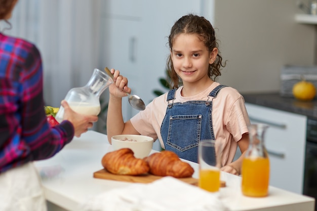 La niña feliz mirando a la cámara con un desayuno saludable junto con la madre en la cocina.