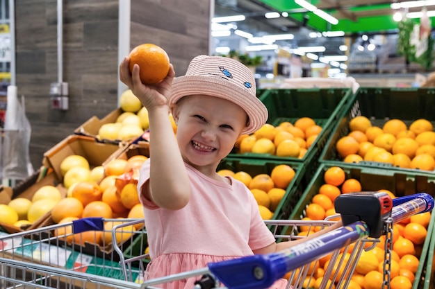 Niña feliz con naranja sentada en un carrito de compras en una tienda de frutas divirtiéndose y mirando a la cámara Un niño lindo comprando frutas frescas al por menor