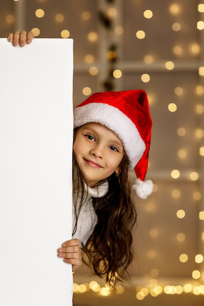 Niña feliz niño con sombrero rojo de santa sosteniendo pancarta de cartón blanco en blanco con espacio vacío