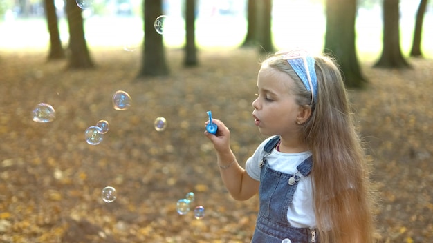 Foto niña feliz niño soplando pompas de jabón afuera en el parque verde. actividades de verano al aire libre para el concepto de niños.