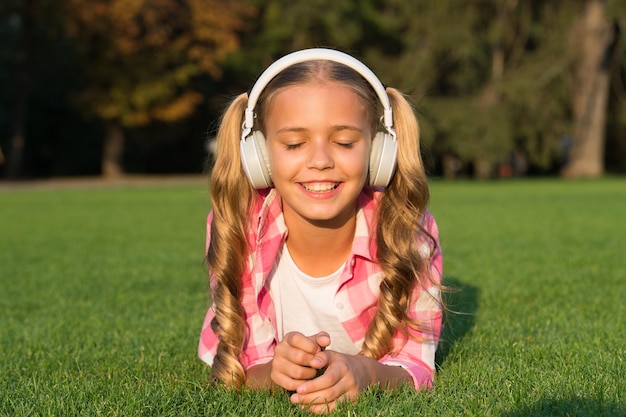 Una niña feliz con los ojos cerrados escucha música en auriculares relajándose en la hierba verde sonido al aire libre de verano soleado