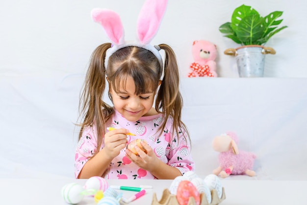 Niña feliz con orejas de conejo pintando el huevo con un bolígrafo de fibra preparándose para el feliz día de Pascua