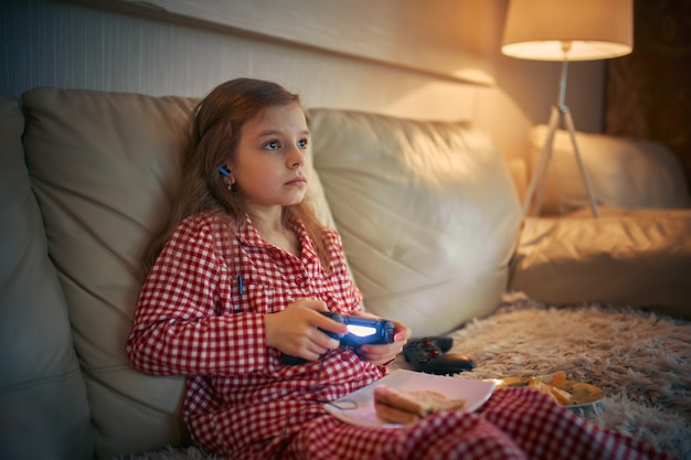 Niña feliz en pajams sentado en el sofá, comiendo pizza y jugando videojuegos con joystick en casa.