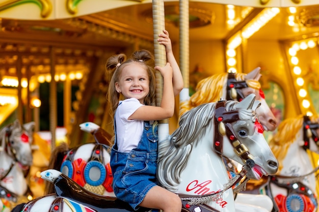 Niña feliz en un parque de atracciones monta un caballo en un carrusel en el verano